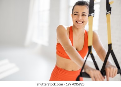 Portrait of a cheerful athletic woman training on suspension straps at white sunny gym. TRX training on fitness straps, happiness and healthy lifestyle