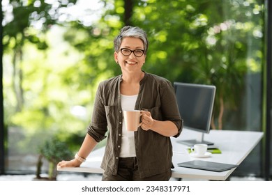 Portrait of cheerful attractive mature businesswoman in smart casual wearing eyeglasses drinking coffee at home office, standing by workdesk with laptop and computer pc on it, smiling at camera - Powered by Shutterstock