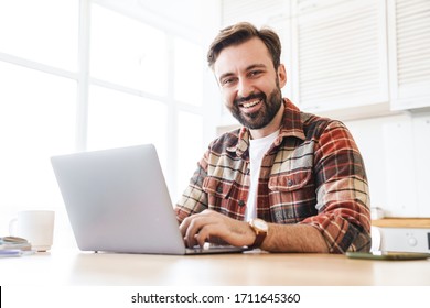 Portrait of cheerful bearded man working with laptop and laughing while sitting at table in home - Powered by Shutterstock