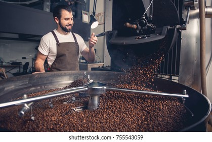 Portrait of cheerful bearded master having job at factory. He controlling brown beans pouring into cooler machine from large coffee roaster. Work concept