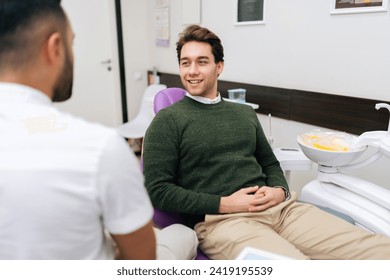 Portrait of cheerful bearded young man patient attentively listening unrecognizable male dentist discusses treatment plan sitting on dental chair in dentistry clinic. Concept of dentistry, teeth care. - Powered by Shutterstock