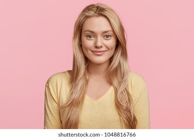 Portrait of cheerful beautiful blonde woman with pleased look, wears yellow casual sweater, models in studio against pink background. Content young female has pleasant smile on face. Horizontal shot
