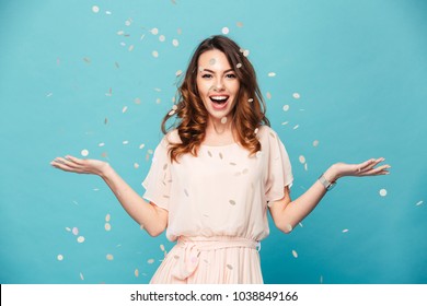 Portrait of a cheerful beautiful girl wearing dress standing standing under confetti rain and celebrating isolated over blue background