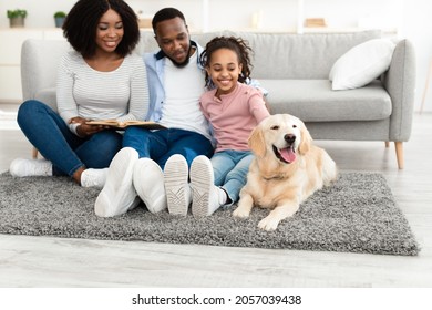 Portrait Of Cheerful Black Mom, Dad And Girl Reading Paper Book Spending Free Time Together Sitting On The Rug Floor Carpet, Smiling Daughter Patting Labrador, Selective Focus On Happy Golden Retiever