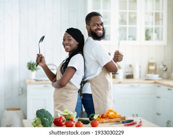 Portrait Of Cheerful Black Spouses Wearing Aprons Posing In Kitchen While Cooking Dinner Together, Happy African American Man And Woman Holding Kitchenware Utensils In Hands And Smiling At Camera