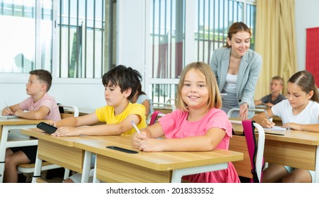 Portrait Of Cheerful Blonde Tween Girl Studying In Classroom, Listening To Schoolteacher And Writing In Notebook