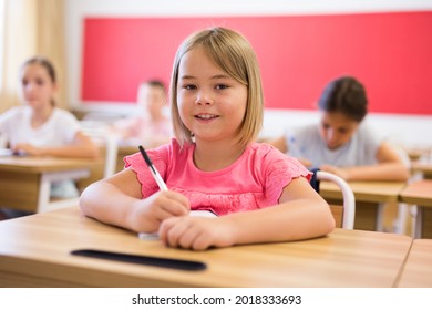 Portrait Of Cheerful Blonde Tween Girl Studying In Classroom, Listening To Schoolteacher And Writing In Notebook