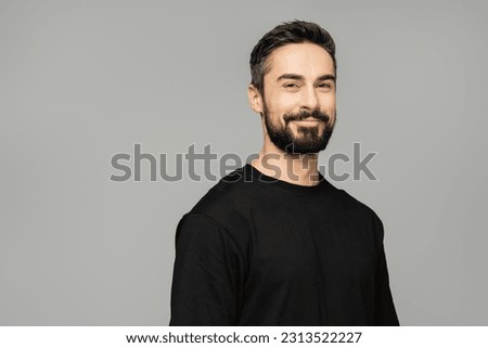 Portrait of cheerful brunette and bearded man in casual black t-shirt smiling at camera while standing isolated on grey, masculine beauty concept, confident and charismatic