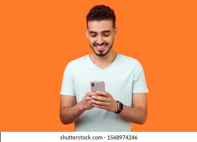 Portrait of cheerful brunette man with beard in white t-shirt using cellphone and smiling, reading good news message, enjoying mobile application. indoor studio shot isolated on orange background - Shutterstock ID 1568974246
