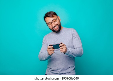 Portrait Of Cheerful Brunette Man With Glasses Wears A Gray Sweater Using Cellphone And Smiling, Reading Good News Message, Enjoying Mobile Application. Indoor Studio Shot, Over Color Background      