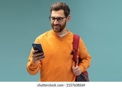 Portrait Of Cheerful Brunette Student Man With Beard Using Cellphone And Smiling, Reading Good News Message, Enjoying Mobile Application. Indoor Studio Shot Isolated On Blue Background 