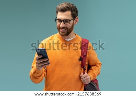 Portrait of cheerful brunette student man with beard using cellphone and smiling, reading good news message, enjoying mobile application. Indoor studio shot isolated on blue background 