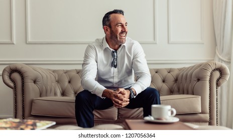 Portrait of cheerful businessman sitting on sofa in hotel room and looking away laughing. CEO staying in luxurious hotel room on business trip. - Powered by Shutterstock