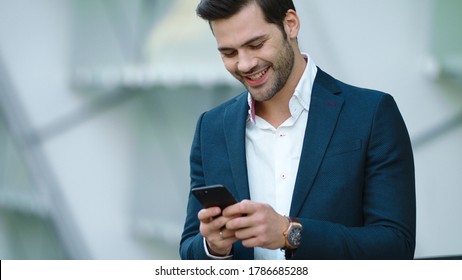 Portrait cheerful businessman using smartphone at modern street. Joyful business man smiling with phone in hand outdoors. Successful businessman talking phone in suit outside
