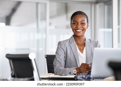 Portrait of a cheerful businesswoman sitting at desk in modern office and looking at camera. Smiling african american executive using laptop while working from office with copy space. - Shutterstock ID 2250909397