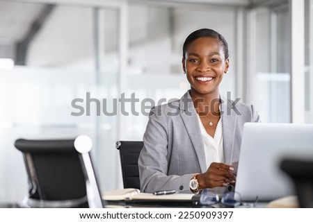Portrait of a cheerful businesswoman sitting at desk in modern office and looking at camera. Smiling african american executive using laptop while working from office with copy space.