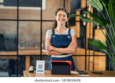 Portrait Of Cheerful Cafe Owner Standing With Arms Crossed And Digital Tablet, Qr Code And Nfc Machine On Table. Successful Woman In Cafeteria Wearing Apron And Looking At Camera. Satisfied Worker.