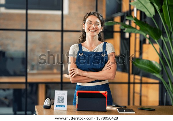 Portrait of cheerful cafe owner standing with\
arms crossed and digital tablet, qr code and nfc machine on table.\
Successful woman in cafeteria wearing apron and looking at camera.\
Satisfied worker.