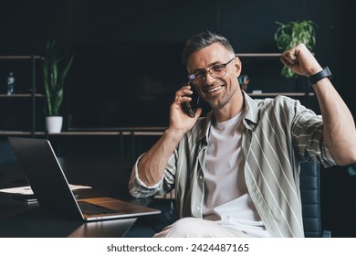 Portrait of cheerful Caucasian businessman in classic eyewear smiling at camera during mobile communication in office with laptop technology, successful male entrepreneur satisfied with received news - Powered by Shutterstock