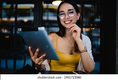 Portrait of cheerful Caucasian female blogger in eyewear smiling at camera during time for online networking via digital tablet, joyful hipster girl with modern touch pad posing in street cafe