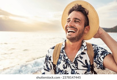 Portrait of cheerful caucasian young man with hat and backpack enjoying sunset at the beach - Laughing guy having fun outside - Well being, healthy life style and traveling concept - Powered by Shutterstock