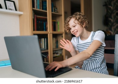 Portrait Of Cheerful Child Girl Sitting At Table Having Video Call, Looking At Laptop Display And Waving Hand To Friends, Family, Parents, Teacher. Preteen Girl Bed Making Videocall Via Laptop 