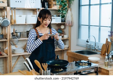 Portrait Of A Cheerful Chinese Girl Using Her Smartphone To Take Pictures Of The Food In The Frying Pan While Cooking In A Rustic Kitchen With Daylight At Home.