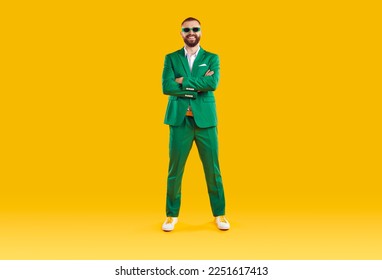 Portrait of cheerful cool and joyful man in green suit and sunglasses of same color. Funny man who celebrates St. Patrick's Day standing with folded hands and smiling at camera on orange background.