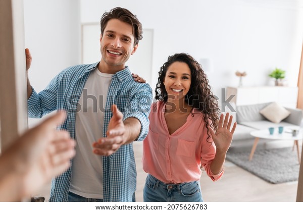 Portrait\
of cheerful couple inviting guests to enter home, happy young guy\
and lady standing in doorway of modern flat, looking out, man\
shaking hands, meeting new neighbors or\
friends