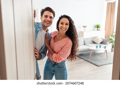 Portrait Of Cheerful Couple Inviting Visitor People To Enter Their Home, Happy Young Guy And Lady Standing In Doorway Of Modern Apartment, Millennial Family Holding Door Looking Out Together