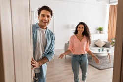 Portrait Of Cheerful Couple Welcoming Inviting Guests To Enter Home, Happy Young Guy And Lady Standing In Doorway Of Modern Flat, Looking Out Together, Waiting For Visitor Friends To Come In