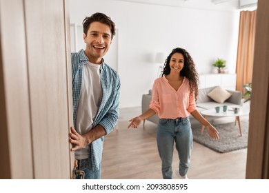 Portrait of cheerful couple welcoming inviting guests to enter home, happy young guy and lady standing in doorway of modern flat, looking out together, waiting for visitor friends to come in