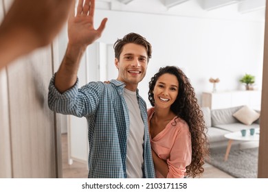 Portrait Of Cheerful Couple Welcoming Inviting Guests To Enter Home, Happy Young Guy And Lady Standing Hugging In Doorway Of Modern Flat, Looking Out Together, Man Giving High Five To Visitor Friend