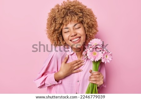 Portrait of cheerful curly haired lady keeps hand on chest glad to receive warm words of congratulation holds bouquet gets her favorite flowers smiles broadly wears shirt isolated over pink background
