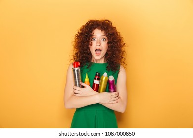 Portrait of a cheerful curly redhead woman holding lots of hair care products isolated over yellow background