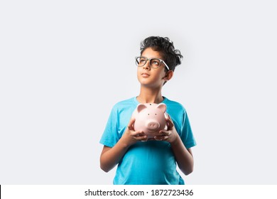 Portrait Of A Cheerful Cute Little Indian Asian Kid Holding Piggy Bank With Books And Looking At Camera Isolated Over White Background