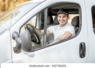 Portrait Of A Cheerful Delivery Driver In Uniform Looking Out The Window Of The White Cargo Van Vahicle, Delivering Goods By Car