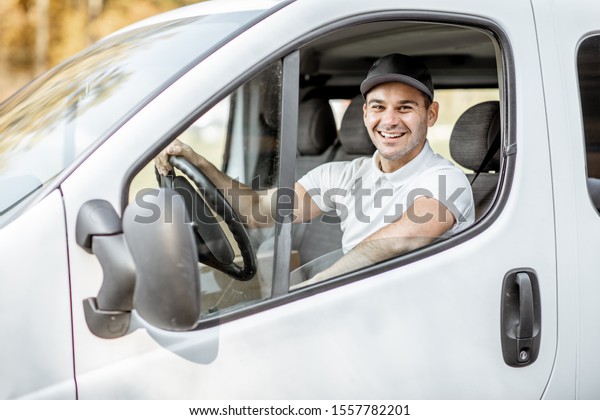 Portrait of a cheerful delivery driver in uniform\
looking out the window of the white cargo van vahicle, delivering\
goods by car