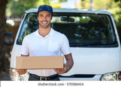Portrait of cheerful delivery person holding cardboard box while standing by van