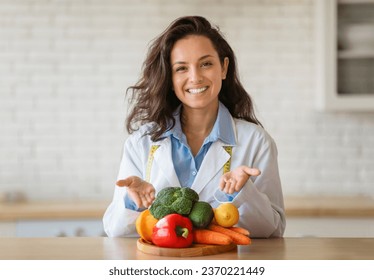 Portrait of cheerful dietitian in lab coat pointing at fresh fruits and vegetables on table, smiling at camera at clinic. Weight loss consultant recommending healthy plant based diet