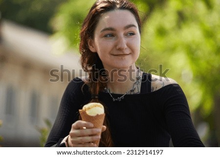 Portrait of a cheerful diverse female with tattoo and lip piercing eating an ice cream in a sunny park. Natural looking female person enjoying a dessert food in summer