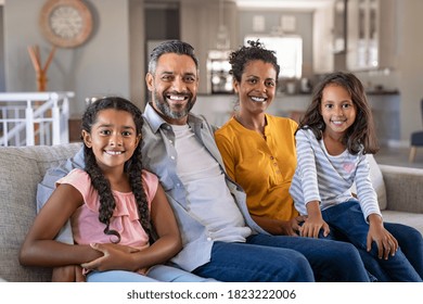 Portrait Of Cheerful Ethnic Family At Home Sitting On Sofa And Looking At Camera. Happy Indian Family With Two Children Relaxing At Home. Mixed Race Parents With Their Daughters In New Home.