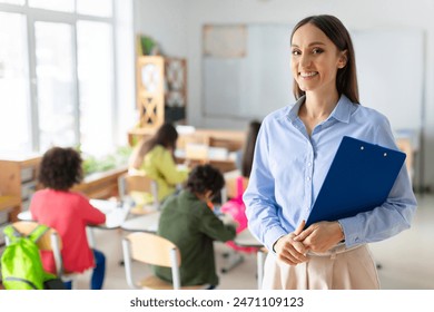Portrait of cheerful European female teacher standing in classroom during lesson with pupils studying on background - Powered by Shutterstock