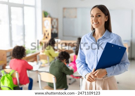 Portrait of cheerful European female teacher standing in classroom during lesson with pupils studying on background