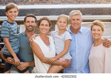 Portrait Of Cheerful Extended Family Standing Outside Their House. Happy Family Standing Outside And Looking At Camera. Parents With Grandparents E Children Enjoying Vacation Together.
