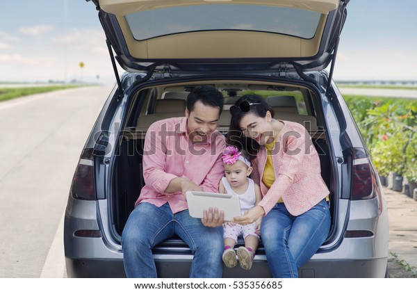 Portrait of cheerful family sitting in the\
car baggage while looking at digital\
tablet