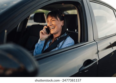 Portrait of a cheerful fashionable young woman sitting on front seat in a car with seat belt on and having a phone conversation. A girl is laughing and talking on her phone while driving an automobile - Powered by Shutterstock