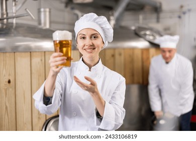 Portrait of cheerful female brewmaster in white uniform presenting glass of fresh beer. - Powered by Shutterstock