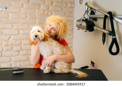 Portrait Of Cheerful Female Groomer Hugging Adorable Curly Labradoodle Dog Before Brushing And Shearing, Preparation To Bathing At Grooming Salon. Woman Pet Hairdresser Doing Professional Care.