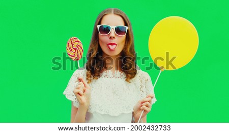Portrait of cheerful funny young woman showing her tongue with lollipop and yellow balloon on green background